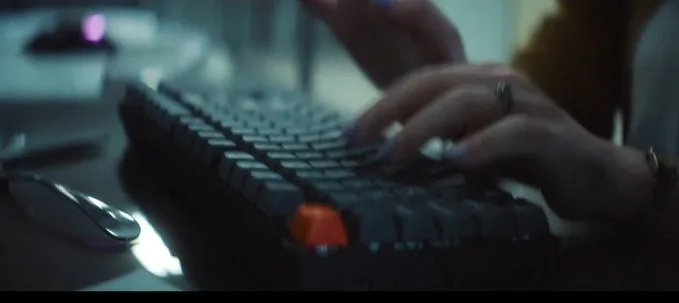 A frame from OpenAI's film — a dark Keychron keyboard with a single orange key, hands typing